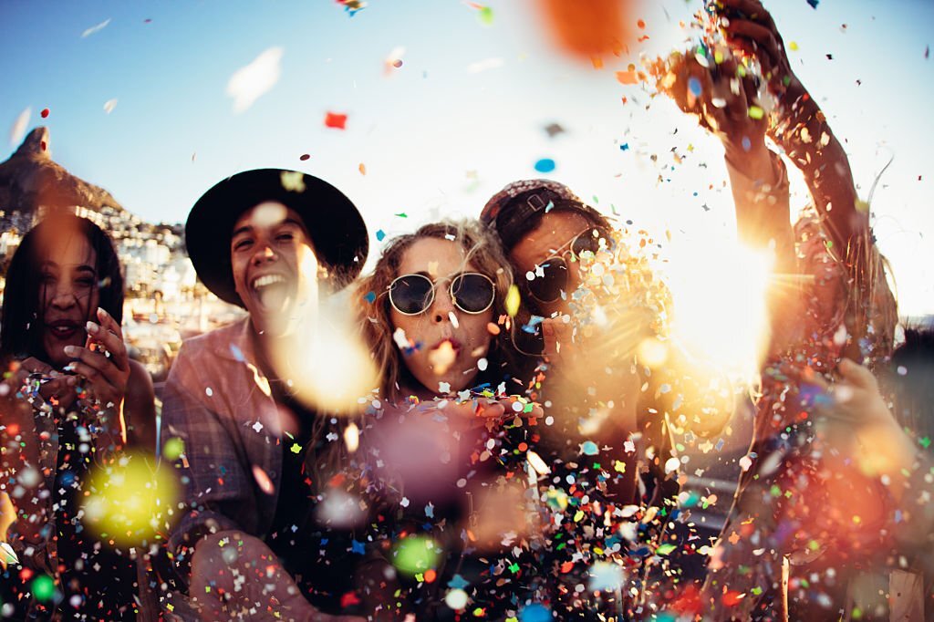 Group of teenager hipster friends partying by blowing and throwing colorful confetti from hands with sunset sun flare